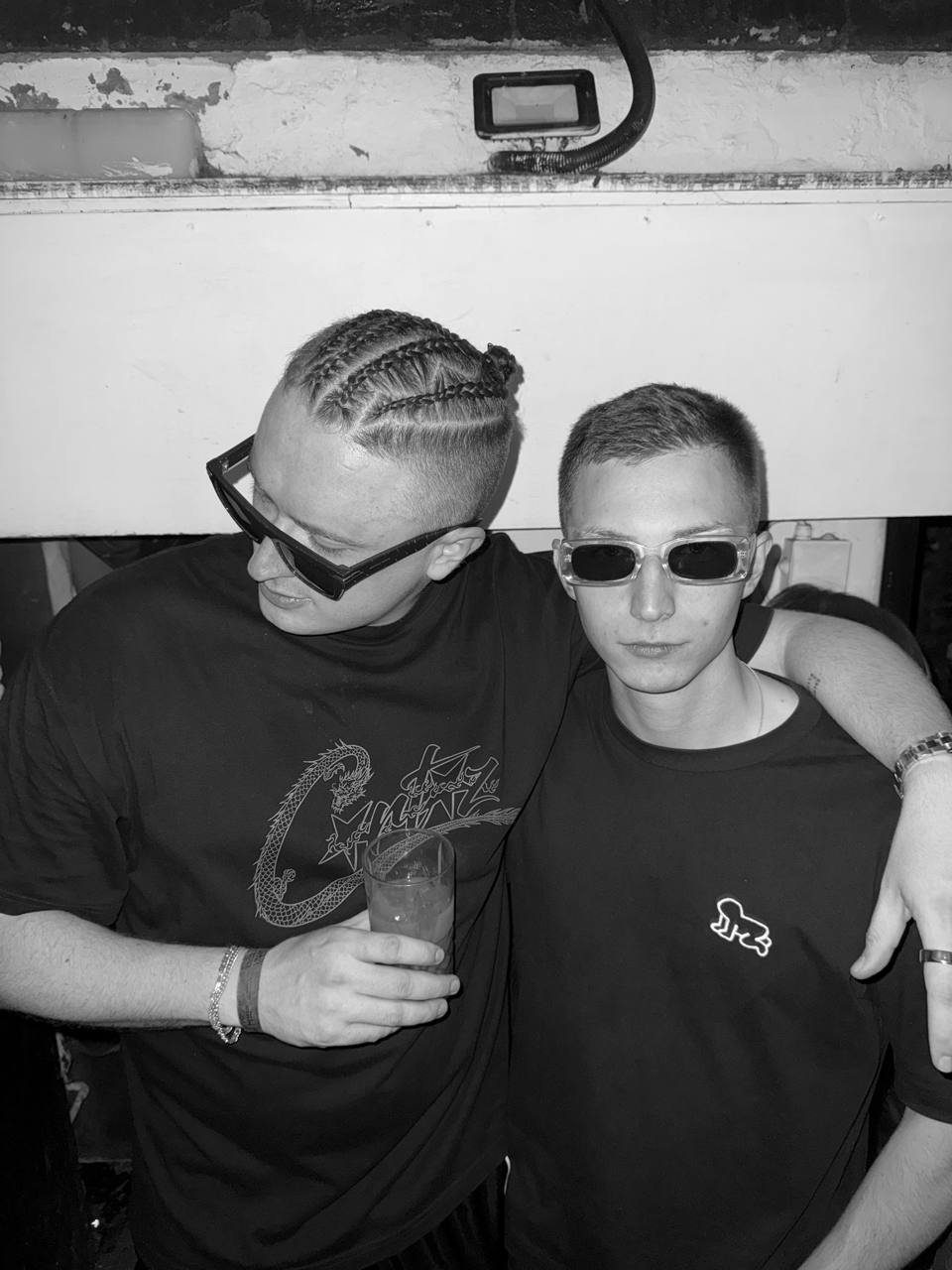 MNEEMO (Yaroslav Gorovoy) & SLAVKO (Viacheslav Zazymko) posing in sunglasses, with one of them holding a glass. Dark background, both wearing black graphic T-shirts, relaxed atmosphere.