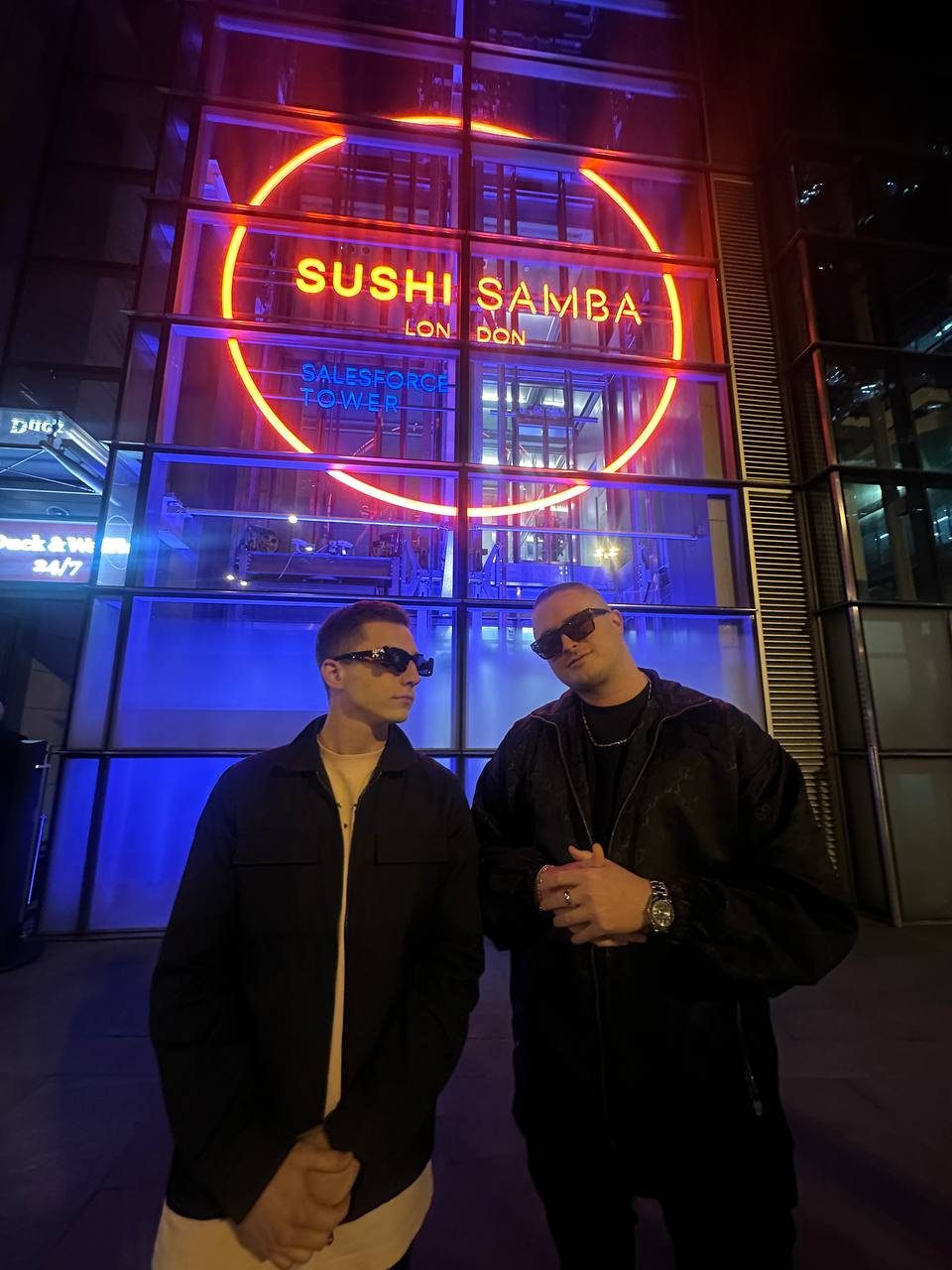 MNEEMO (Yaroslav Gorovoy) & SLAVKO (Viacheslav Zazymko) wearing dark jackets and sunglasses, standing in front of the neon SUSHI SAMBA sign in London. The background is lit with red and blue lights.