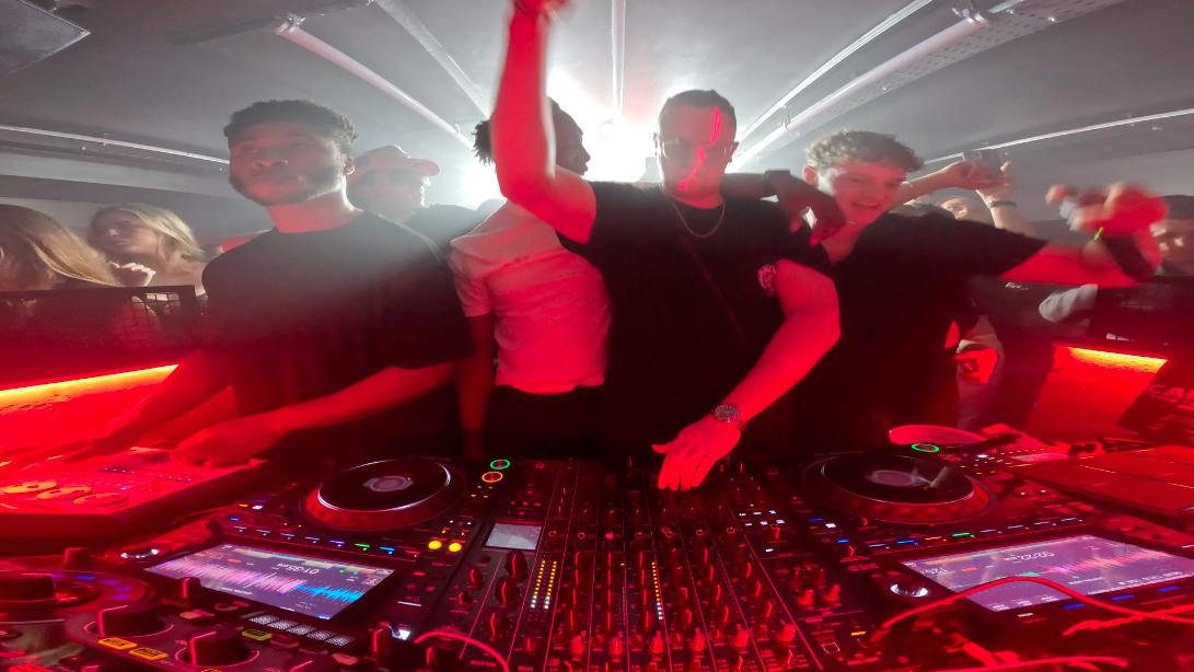 MNEEMO (Yaroslav Gorovoy) & SLAVKO (Viacheslav Zazymko) behind the DJ booth in a club, under atmospheric red lighting, while people dance and clap, creating a vivid and energetic atmosphere.