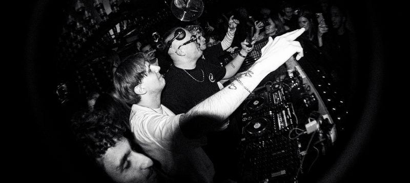 Dancefloor scene, MNEEMO (Yaroslav Gorovoy) wearing headphones behind the decks, in black and white, with a crowd filming on their phones. Energetic atmosphere.