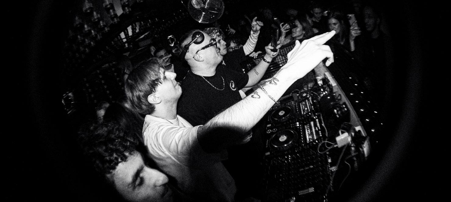 Dancefloor scene, MNEEMO (Yaroslav Gorovoy) wearing headphones behind the decks, in black and white, with a crowd filming on their phones. Energetic atmosphere.