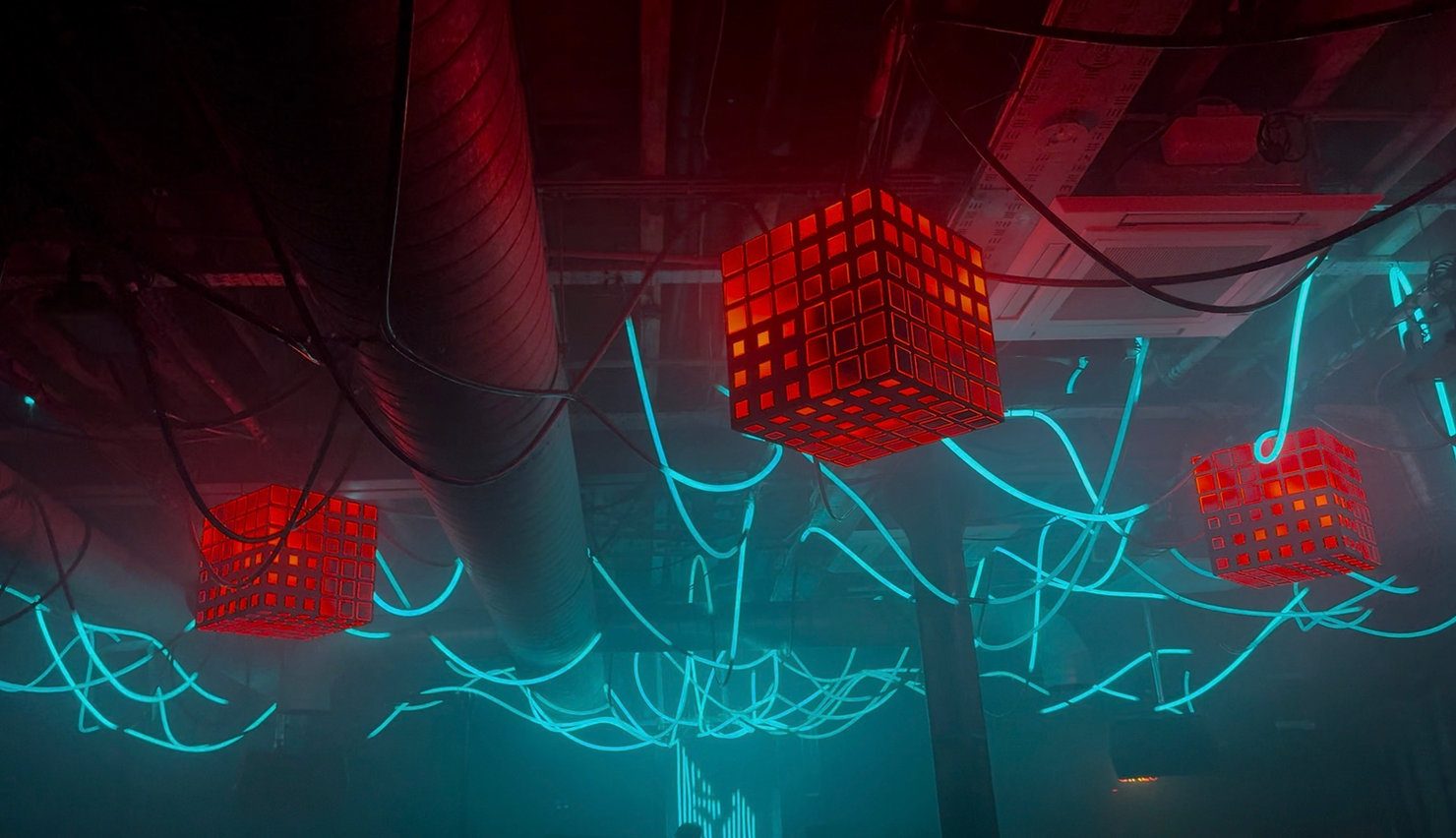 Red cubes hang from the ceiling among blue neon tubes in a dark room, creating a futuristic atmosphere. Shot during MNEEMO’s (Yaroslav Gorovoy) interview with Kirk Allen at the opening of XOYO London.