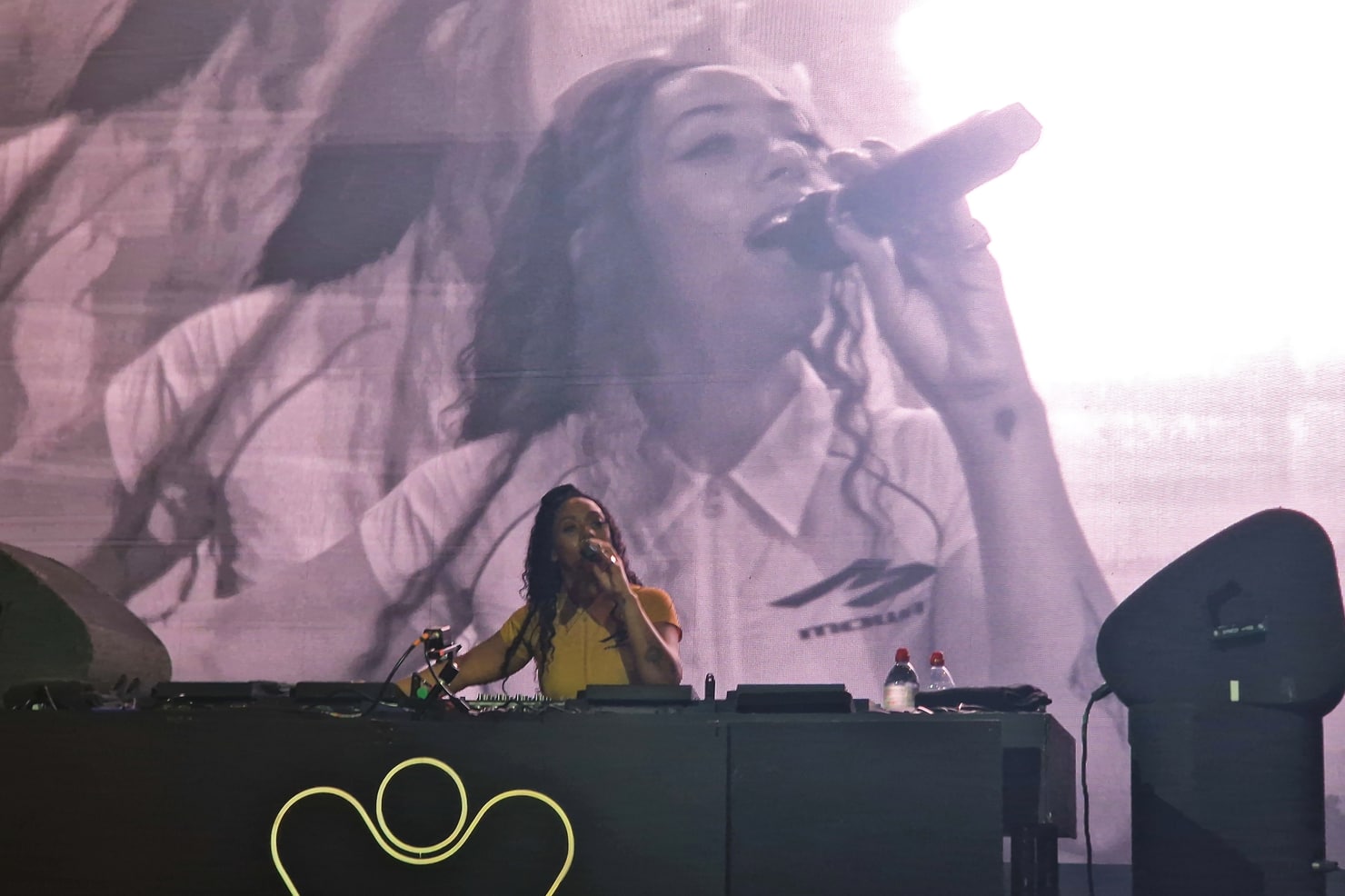 Nia Archives behind the decks with a microphone at a live set in Amsterdam, her face on the big screen behind her