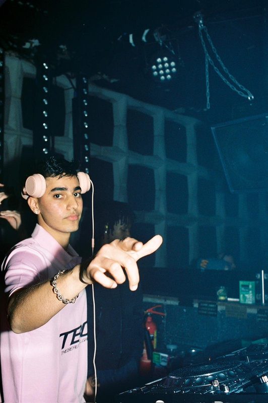 OMAR+ in a pink T-shirt and headphones behind the DJ booth in a club. He is pointing forward, with a darkened background and a glowing ceiling visible.