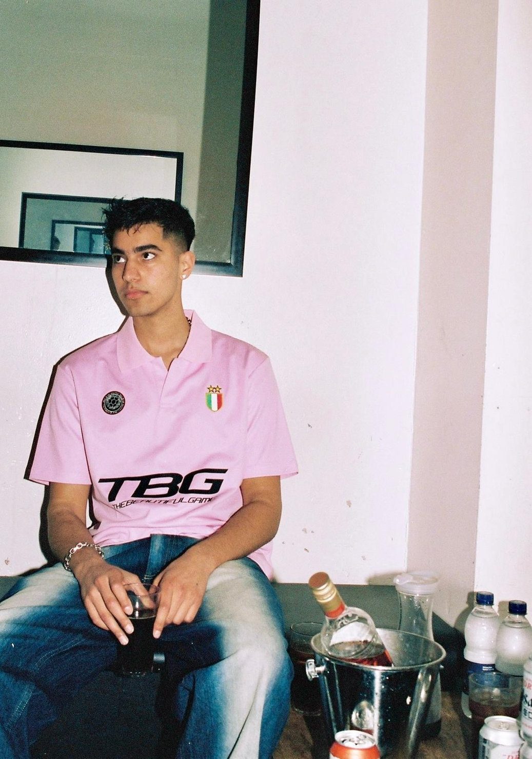 OMAR+ in a pink T-shirt with logos, sitting with a drink in hand. In the background, there is a mirror and a table with bottles. Calm atmosphere.