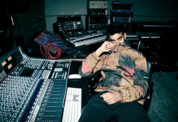 OMAR+ in a camouflage jacket, sitting at a mixing desk in the studio, holding his collar. Equipment and a keyboard are visible in the background.