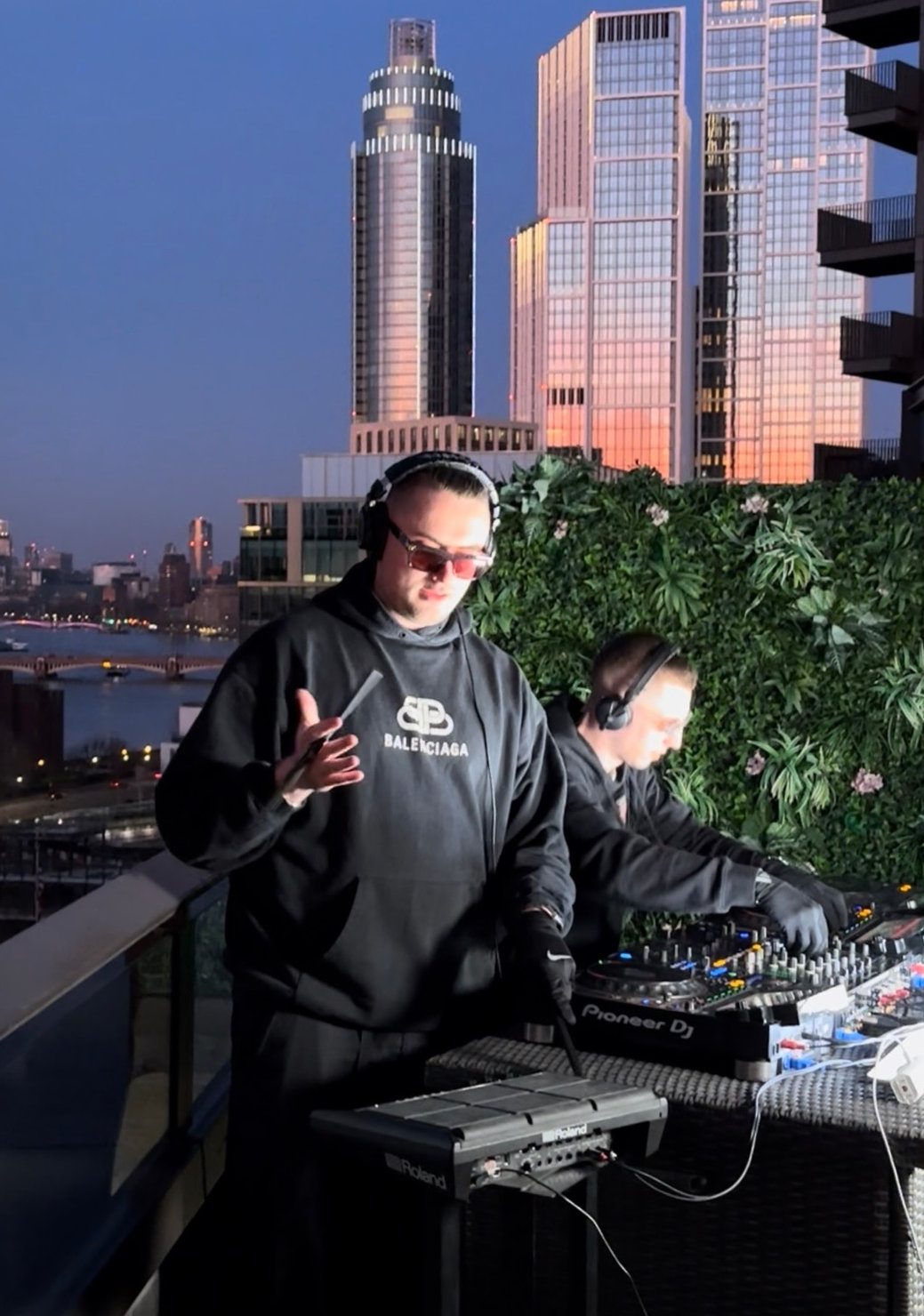 MNEEMO (Yaroslav Gorovoy) & SLAVKO (Viacheslav Zazymko) on a balcony at sunset. One wears a Balenciaga hoodie, headphones, and shades. City skyline and greenery backdrop.