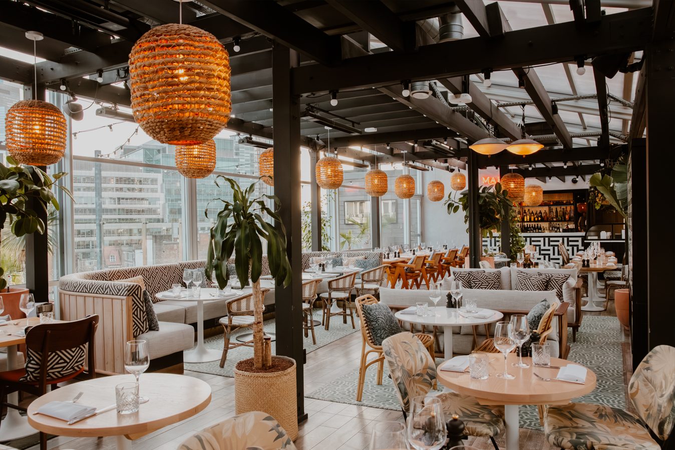 45 London on 45 Curtain Road. Chic restaurant interior with woven pendant lights, patterned cushions, and plants. Wooden tables set for dining, surrounded by floor-to-ceiling windows.