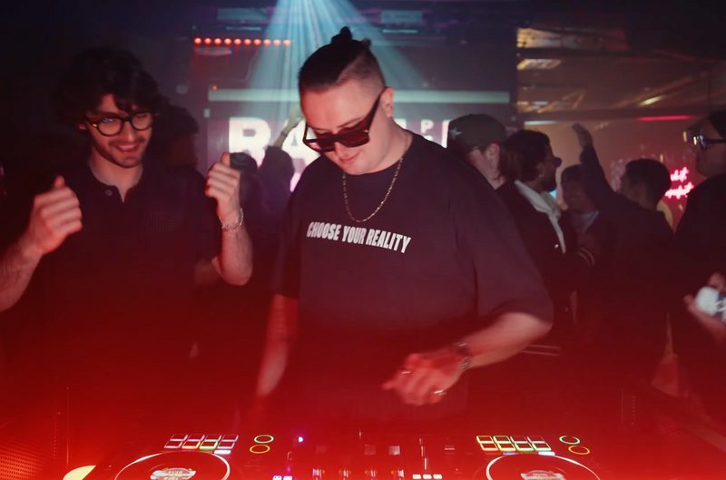 MNEEMO (Yaroslav Gorovoy) in a club, wearing glasses and a “Choose Your Reality” T-shirt behind the DJ booth, with bright lights and a lively atmosphere.