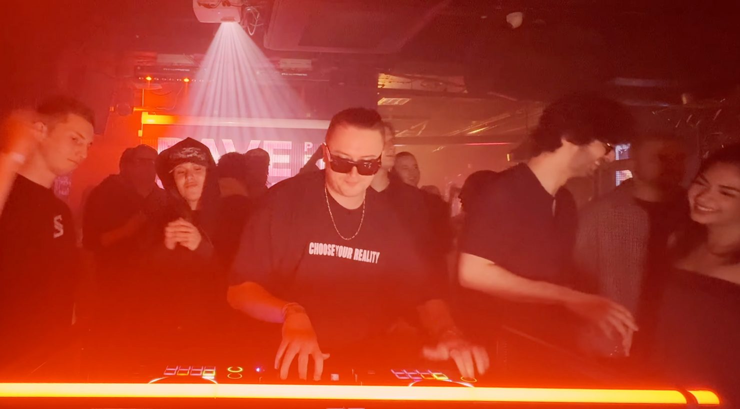 Диджей MNEEMO (Yaroslav Gorovoy) wearing a black T-shirt with the words “CHOOSE YOUR REALITY”, performing on the decks. A crowd is gathered around him under red lighting in a club.