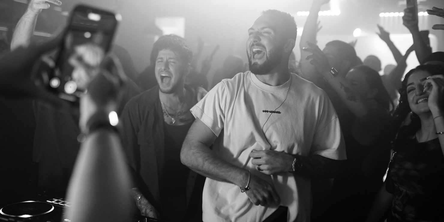 People at a party, laughing loudly and dancing. In the foreground: Mike Jasom & D.X.D. Joyful atmosphere, with smoke and lighting in the background.