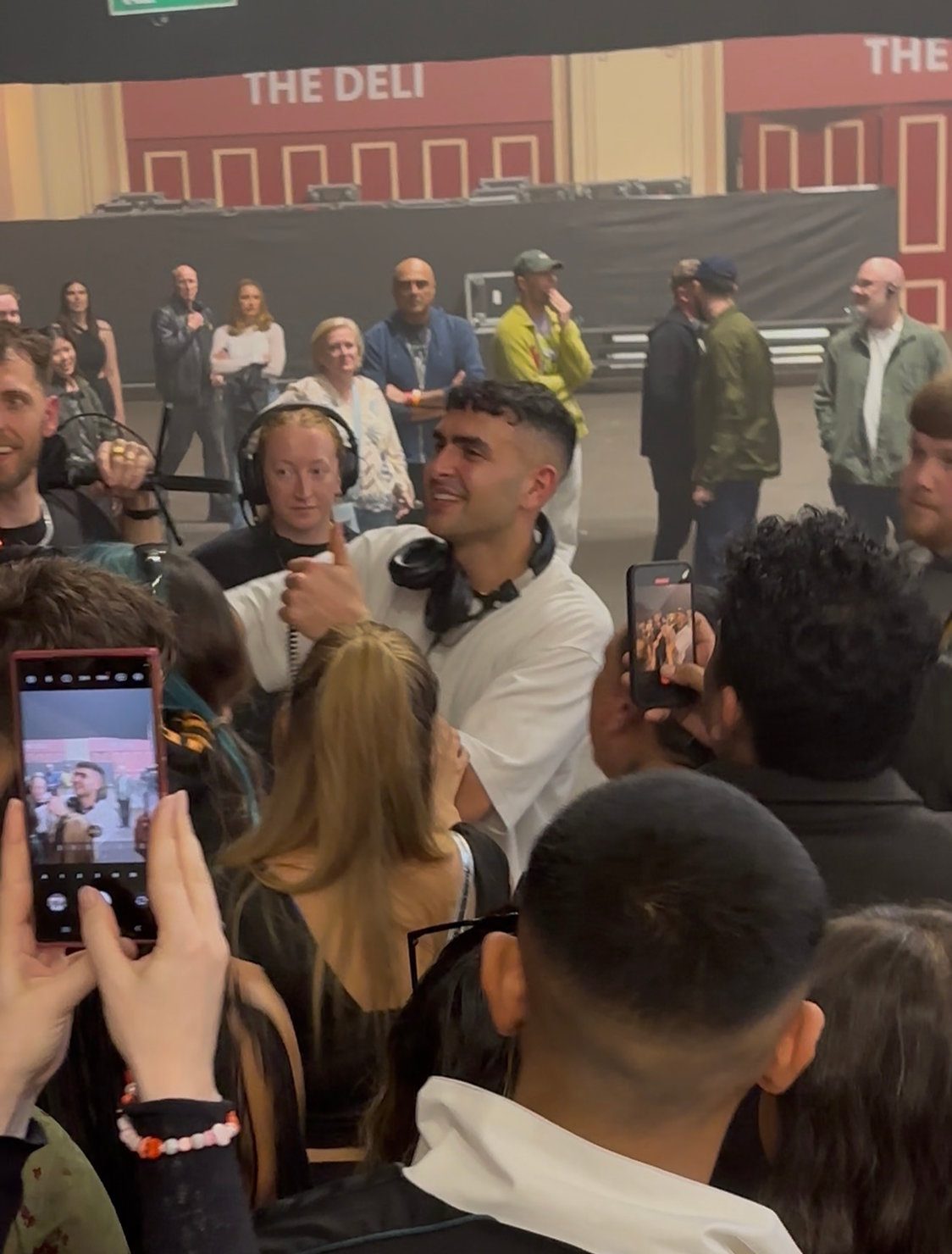 Sammy Virji at Alexandra Palace surrounded by fans after his show in white shirt holds microphone, gestures thumbs up in crowded room. People take photos. Background has "THE DELI" sign. Energetic mood. 18 April 2026
