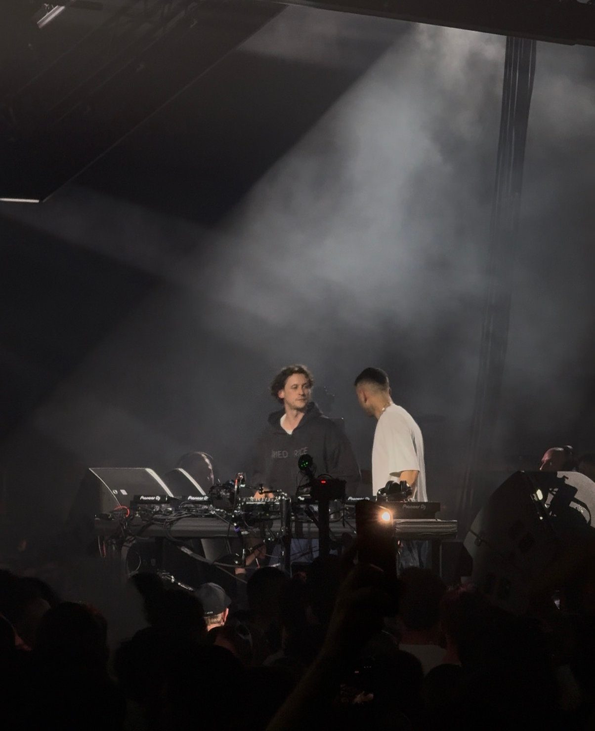 Sammy Virji & Barry Can't Swim at Alexandra Palace performing on stage with a mixer and laptop, surrounded by a smoky atmosphere and crowd. One wears a black hoodie; the other a white shirt. 18 April 2026.