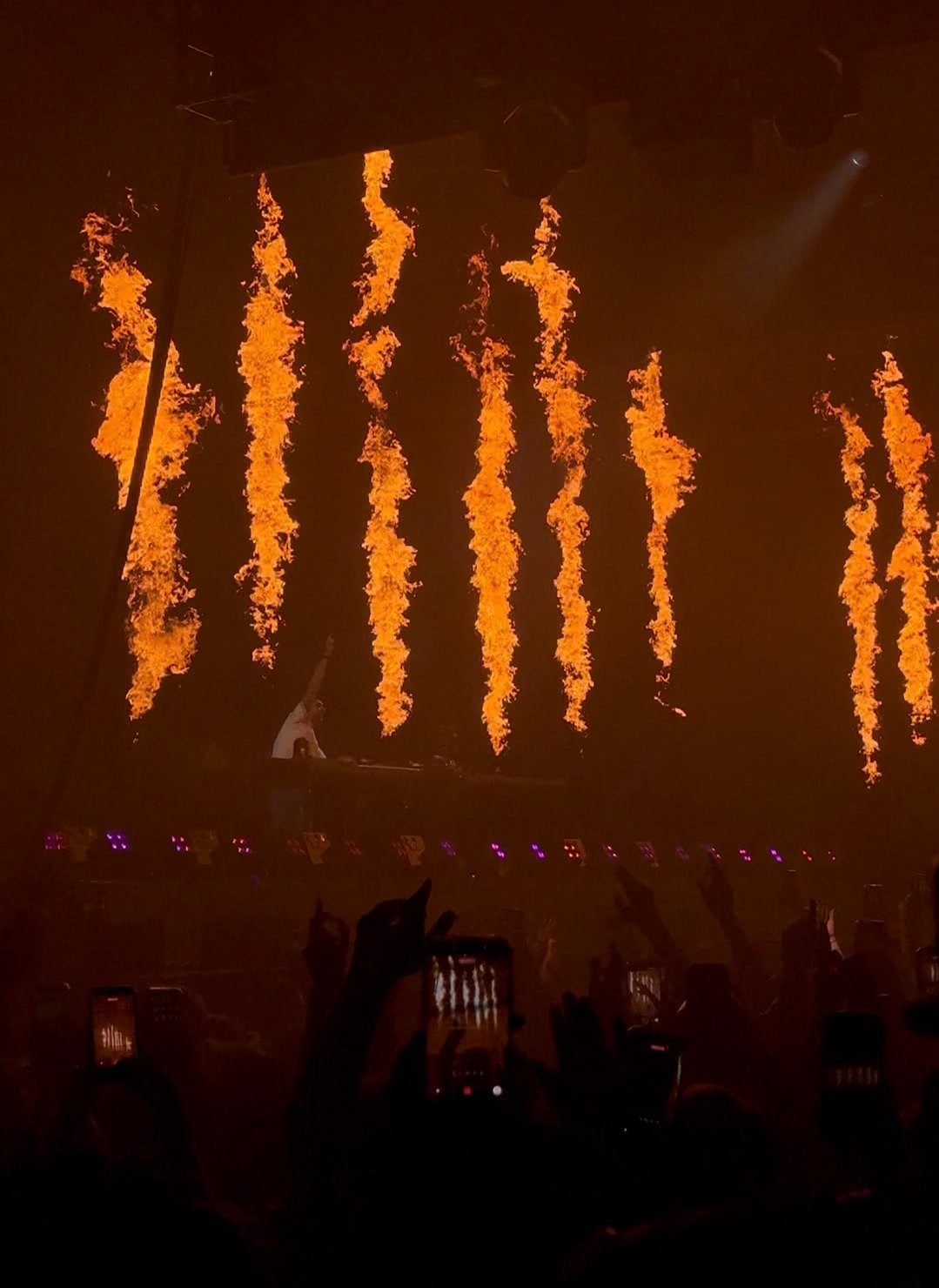 Sammy Virji DJing at Alexandra Palace on stage with hands raised behind flaming pyrotechnics. Crowd with raised hands and phones capturing the fiery display in a dimly lit venue. 18 April 2026.