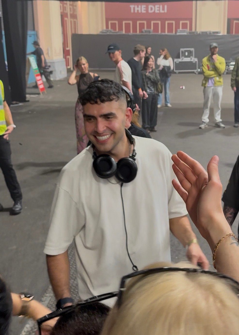 Sammy Virji after his show at Alexandra Palace 18 April 2026 wearing headphones smiles in a lively indoor setting. People stand in the background. "THE DELI" sign visible above.