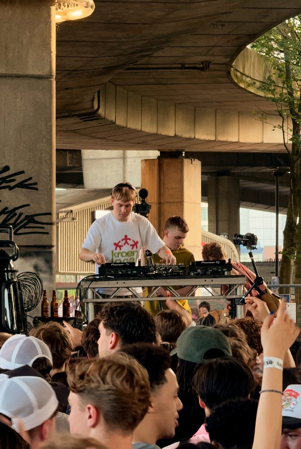 Silva Bumpa DJing at his HYPA EP launch in London, standing behind Pioneer DJ decks on a pickup-style setup with the crowd filming and sunlight breaking through the trees.
