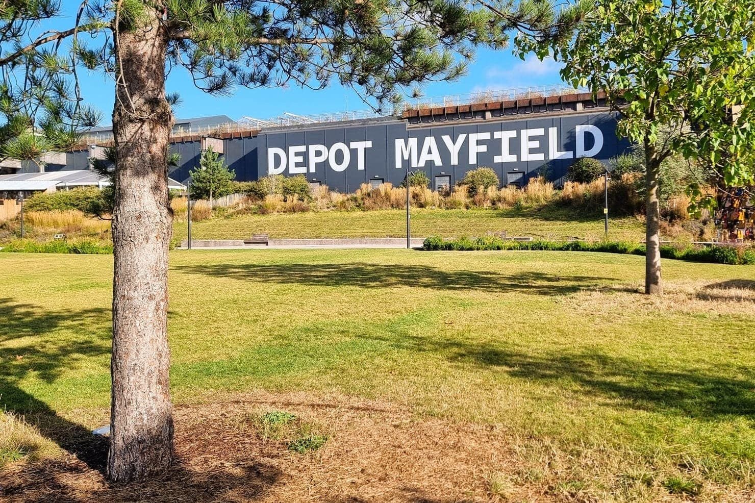 The exterior of Depot Mayfield in Manchester in daylight, the words Depot Mayfield painted in white on the dark wall above a park