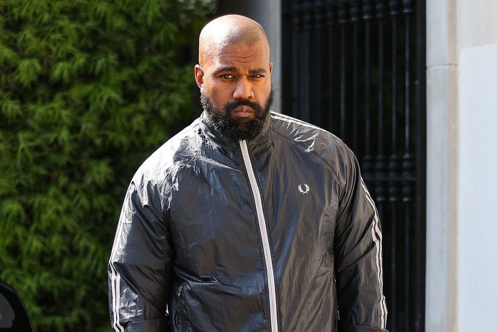 Kanye West (Ye) in a dark jacket, walking with greenery and a building in the background. Serious expression. A logo is visible on the left side of the jacket.