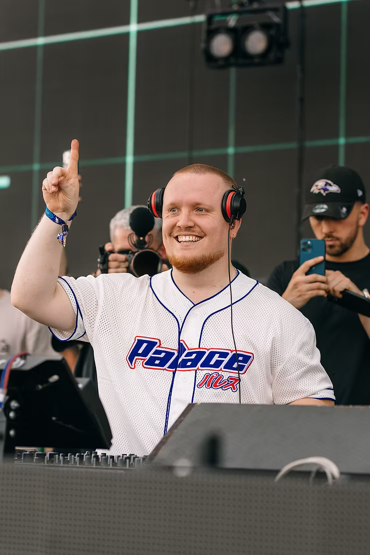 Chris Stussy DJing live in a Palace baseball jersey and headphones, one finger raised mid-set as photographers film from the crowd